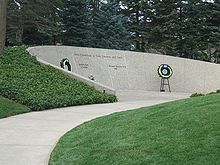 An arching stone wall is set into a small grass-covered hillside. The wall is engraved thus: "Lives Committed to God, Country, and Love", with two names engraved underneath, reading "Gerald R. Ford, 1913–2006" and "Elizabeth Bloomer Ford, 1919–2011 ". Two flowered wreaths are placed beside the names on stands.