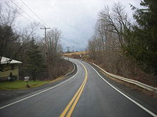 Ground-level view of a two-lane road passing through a rural area, with only one home visible on the left-hand side. Wooded terrain opens to an a field as the highway curves towards the left.