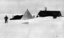  On the right is a long low hut with two chimneys, surrounded by snow drifts. In the centre is a pointed tent and, to the left, a covered mound of stores. Several men are visible, as is a seal.