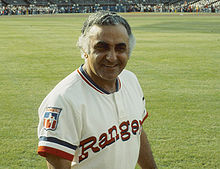 A man with gray hair wearing a white baseball jersey with "Rangers" written across the chest