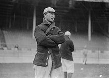 A man wearing a dark-colored coat and baseball cap with his arms crossed stands on a baseball field; two similarly dressed men are behind him.