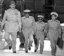Four men in military uniform, wearing buttoned jackets, wide-rimmed hats, black shoes, and carrying briefcases. They are ascending the stairs of a building.
