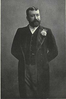 G. R. Sims, a dapper paunchy gentleman with a black beard and hair wearing smart clothes and a buttonhole