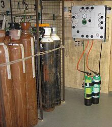A panel on the wall is connected to diving cylinders by hoses. Nearby are several much larger cylinders, some painted brown and others black