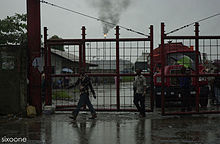 A picture of gates of the Port Harcourt Refining Company Ltd. with an oil refinery's fire seen in the background in rainy weather.