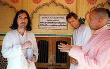Three men in their early fifties, one to the left, wearing a white robe and holding a bottle of water with both hands, and two to the right, one in a white robe and one in a pink robe. On the wall behind them is something written in Sanskrit, both in Roman characters and in Sanskrit characters.