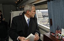 George W. Bush wearing a suit, tie, scarf and overcoat while looking out the window from a skyward vehicle.