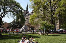 Many students mingle in the background while a group sit in the foreground on a grass lawn. The large stone clocktower is seen above the trees on the lawn.