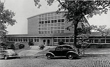 A black-and-white photograph of a one-story building with windows all along it and a multi-story portion of the building set further back from the road. In front of the building, there are two cars parked on a brick-paved road.