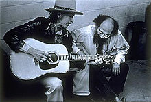 Dylan, wearing a hat and leather coat, plays guitar and sings, seated. Crouched next to him is a bearded man, listening to him with head bent.
