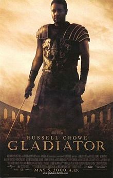 A man standing at the center of the image is wearing armor and is holding a sword in his right hand. In the background is the top of the Colosseum with a barely visible crowd standing in it. The poster includes the film's title, cast credits, and release date.