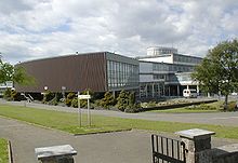 A contemporary school building constructed with glass, concrete and steel with white mosiac tiles. Large brown cladding is used on one side of the assembly hall. A cylindrical glass belvedere feature is used on the roof
