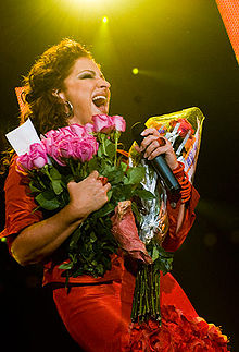A woman stands under a spotlight. She holds several bouquets of flowers and a microphone.