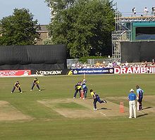 A cricket match with fielders and batmen wearing coloured kit. A bowler delivers a ball to one of the batsman. Some of the crowd can be seen behind advertising hoardings and in front of trees and a scaffold construction.