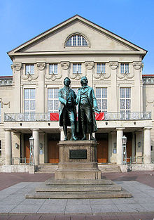 Photograph of a large bronze statue of two men standing side-by-side and facing forward. The statue is on a stone pedestal, which has a plaque that reads "Dem Dichterpaar/Goethe und Schiller/das Vaterland". Behind the monument there is a large, 3-storey building with an elaborate stone facade.