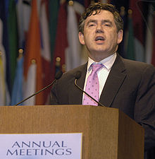 Gordon Brown standing at a podium. Text on the podium states "ANNUAL MEETINGS". A number of flags hang in the background