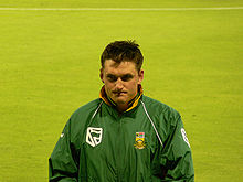 A white man with short dark hair in a green all-weather jacket.  He is standing in front of a large expanse of grass.
