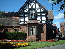 A two-storey house, the lower storey being in sandstone and the upper timber framed and gabled. To the right of the house are the entry gates to the park and in front of it are formal flower beds.