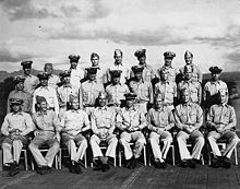 Twenty-eight Sailors in the uniform of the United States Navy pose on the deck of a World War Two-era Aircraft Carrier.
