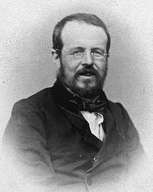 A black and white, half-length portrait of an middle-aged bearded man wearing a dark suit, white shirt, a bow tie and glasses.