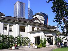 A two-storey neo-classical building showing Japanese architectural influences, with a central two-storey tower. In the foreground is a garden and tennis court and in the background are skyscrapers.