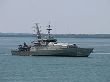 A grey warship underway. Land is visible in the background.