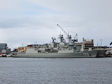 Colour photo of two grey-painted warships moored alongside a wharf. A large crane and several buildings are visible behind the ships.