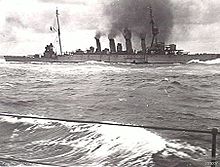 A large four-stacked warship billowing thick black smoke and moving through moderate seas