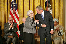 A color photograph of Harper Lee smiling and speaking to President George W. Bush while other seated Medal of Freedom recipients look on