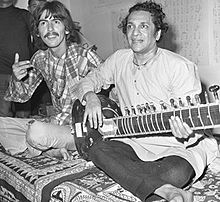 Black-and-white picture of two men, one, in the foreground to the right, in his mid-forties, and the other, in the background to the left, in his mid-twenties. Both are sitting cross-legged on rugs, and the man on the right holds a sitar.