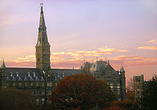 A large Gothic style stone building dominated by a tall clocktower with a pinkish sunset behind it.