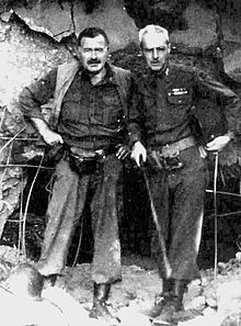 a dark haired man with mustache dressed in army boots, tucked in muddy pants, shirt and vest, with a light haired man dressed in army clothing standing in front of rubble