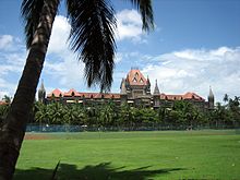 A brown building with a central tower and sloping roofs surrounded by trees. A grassy ground and a coconut tree are in front of it.