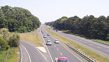 A highway passes beneath the camera and continues straight into the horizon. It is surrounded by forests on either side and contains no guardrail to separate opposite flows of traffic.