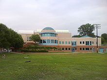A beige building with a large windowed rotunda stands before a large lawn. The sky is overcast.
