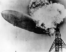 A black and white photograph of an airship near a mooring mast exploding at the rear.