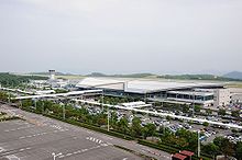 An airport terminal building with an air traffic control tower on the left and a carpark on the foreground of the building