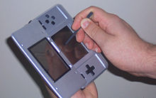 A person holds a handheld video game console in one hand and a writing tool in the other.