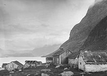 Small wooden huts with pitched roofs, some with no windows, huddle under a steep cliff on a fjord.