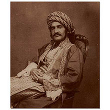 Sepia photograph of a man in 19th century Middle Eastern dress, with a large moustache, reclining in a chair with his hands crossed across his lap