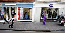 A man in a purple jumpsuit is at the right of the image walking down a street. On the left is a man dressed as a police officer following him. At the far right is a man seated on the back of a golf cart filming them. Storefronts can be seen in the background.