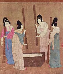 A painted image of four Chinese women wearing colorful silk robes, their hair tied up into buns, standing around a small wooden block with silk laid on top while holding large whisks which they use to beat the silk.