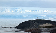  A memorial cross, with a person standing to its left, can be seen distantly, on a headland protruding to the sea, with a backdrop of icy mountains