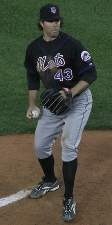A man wearing gray pants, a black baseball jersey with "Mets" written across the chest in blue letters, and a black cap prepares to throw a baseball