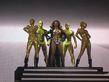 A brunette woman is standing in front of some stairs. She wears a super heroine costume, compound of a golden bra with red diamonds, silver pants, a long black coat and glasses. Behind her five people, dressed like golden mannequins, are standing in different positions.