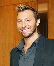 Young man with short blonde-brown hair wearing a dark open necked shirt and suit, not cleanly shaven, smiling broadly. The background is a wooden wall.