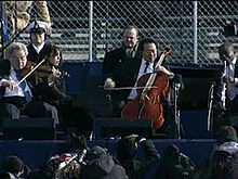 Four male musicians playing a variety of instruments outdoors in front of a chain link fence