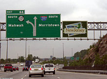 A multilane freeway approaching an interchange with a sign display over the road. The left sign reads exit 66 south Route 17 Mahwah left two lanes exit Interstate 287 south Morristown right two lanes straight and the right sign says Welcome to New Jersey