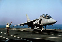 Front view of two-seat grey jet fighter on aircraft carrier deck. A directive personnel is close-by.