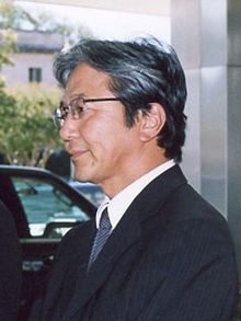 Profile of a middle-aged Japanese man from the chest up wearing a dark suit, blue tie, and glasses. His hair is greying to a silver.